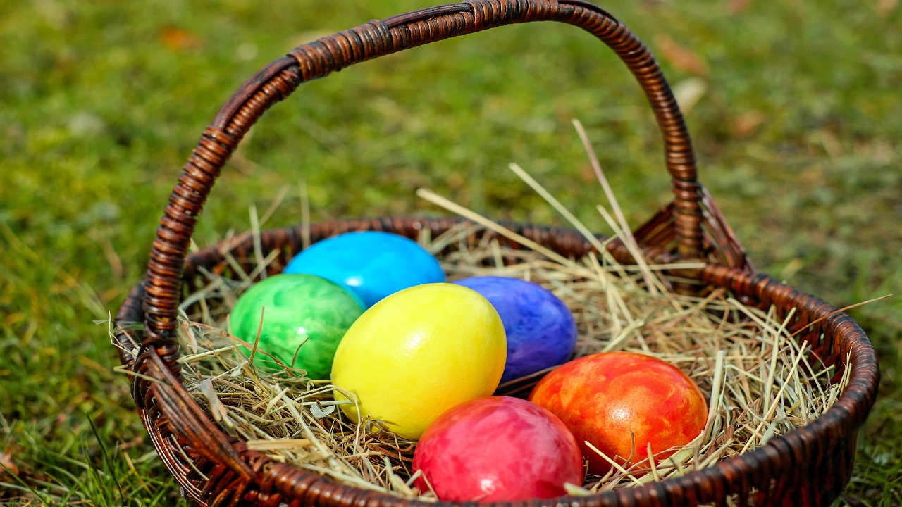 Na trawie postawiony został brązowy koszyk wiklinowy wypełniony sianem, w którym znajdują się kolorowe jajka wielkanocne.